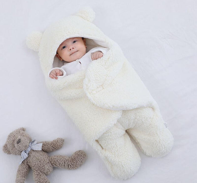 Baby wrapped in a cozy cream quilted sleeping bag with bear ears, beside a fluffy teddy bear.