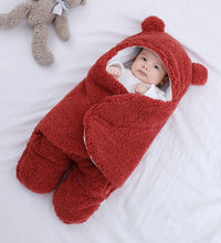 Baby in red quilted sleeping bag with bear ears, resting comfortably on a blanket beside a teddy bear.