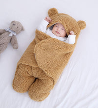Baby wrapped in a cozy brown quilted sleeping bag with bear ears, next to a teddy bear on a white surface.